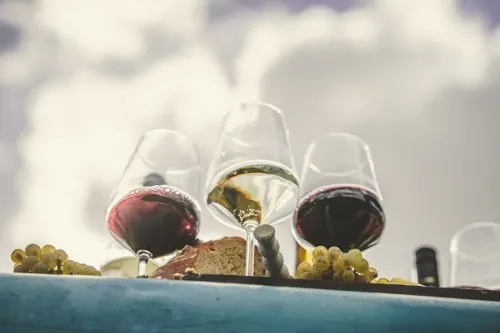 Drei Gläser Wein vor bewölktem Himmel, daneben Trauben und Brot